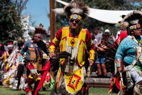 Elder Native American dancer in bright yellow and red regalia leading a traditional procession at Morning Star American Indian Village in Cheyenne.